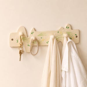 Puede incluir: Un perchero de pared color crema con cuatro ganchos, que presenta un diseño pintado de hojas verdes y pequeñas flores blancas. El perchero sostiene un llavero dorado, una goma para el pelo beige, una bufanda clara y una camisa blanca.