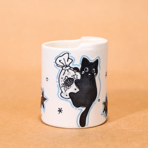May include: White ceramic mug featuring a black cat illustration carrying a bag of goldfish. The mug has blue accents and star details. The design is hand-drawn and whimsical.