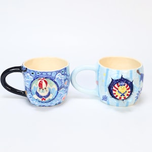 Puede incluir: Dos tazas de cerámica con motivos náuticos. La taza izquierda tiene un asa negra y un relieve de una foca con un gorro de rayas rojas y blancas. La taza derecha tiene un asa azul claro y un relieve de un pollito con traje de marinero.