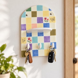 May include: An arched wall-mounted organizer featuring a multicolored checkerboard pattern in shades of blue, yellow, green, purple, and pink. It includes a small white shelf, three white hooks, and decorative butterfly and flower illustrations.