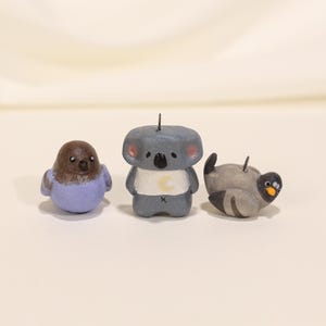 Può includere: Tre piccole statuette di animali fatte a mano: un uccello con la testa marrone e il corpo viola, un koala grigio con una camicia bianca e un piccione grigio. Ogni statuetta ha un piccolo spillo di metallo sopra.