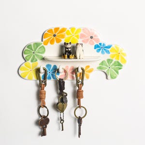 May include: A ceramic cloud-shaped key holder with a floral design in yellow, orange, pink, blue, and green. It features a small shelf with two cat figurines and three keychains with leather straps and metal hardware.