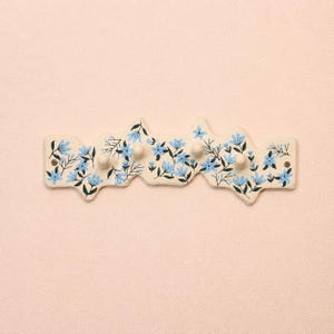 May include: A cream-coloured wall-mounted coat rack with an irregular, wavy edge. The surface is decorated with a hand-painted pattern of light blue flowers and dark green stems. It features four small, rounded knobs and two visible screw holes.
