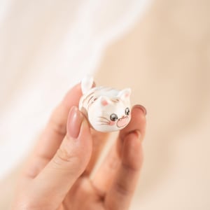 May include: A small, ceramic cat figurine with a white base and brown stripes. The cat has large, expressive eyes, pink cheeks, and a small, upturned tail. The figurine is held in a hand.