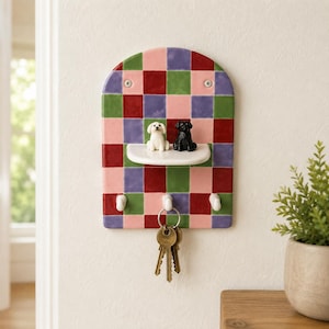 Op de afbeelding: Een wand-sleutelhouder met een kleurrijk ruitpatroon in rood, groen, paars en roze. Het bevat een kleine witte plank met twee miniatuur hondjes, een witte en een zwarte, en drie witte haken met metalen sleutels eraan.