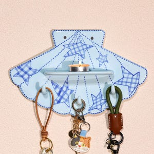 May include: A light blue ceramic wall-mounted key holder shaped like a seashell, decorated with blue plaid stars. It has a small shelf with a lit candle and three hooks holding keychains with leather and fabric accents.