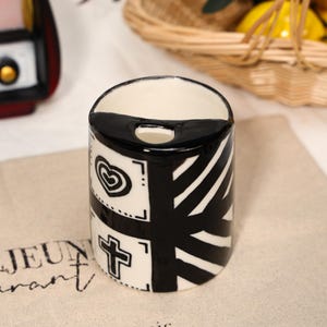 Può includere: Un contenitore cilindrico in ceramica con un design geometrico in bianco e nero. Il design include un cuore, una croce e strisce diagonali. Il bordo superiore è nero e l'interno è bianco. Il contenitore è su una superficie beige.