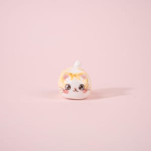 May include: A small, ceramic cat figurine with a rounded body and a cute, cartoonish design. The cat is primarily white with yellow and orange markings, pink cheeks, and large, expressive eyes. The figurine has a small tail and is set against a soft pink background.
