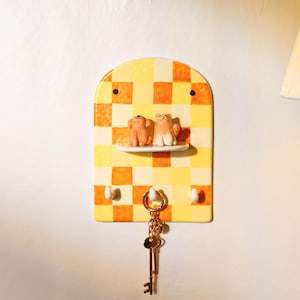 Puede incluir: Un soporte de pared para llaves de cerámica con parte superior arqueada y un patrón de cuadros amarillos y naranjas. Incluye un pequeño estante horizontal con dos figuras de animales en miniatura y tres ganchos blancos, con un llavero dorado.