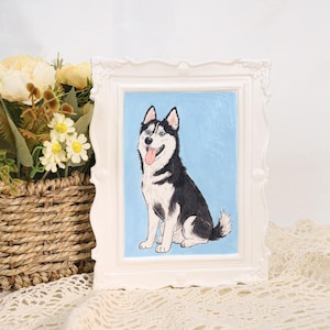 May include: A framed painting of a Siberian Husky dog. The dog is depicted with black and white fur, blue eyes, and a pink tongue. The painting is set against a light blue background and framed in a white, ornate frame. The artwork is displayed on a lace-covered surface.