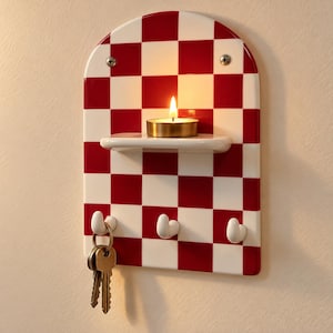 May include: A wall-mounted arched plaque featuring a red and white chequered pattern. It includes a small horizontal shelf holding a lit tea light candle and three white hooks at the bottom, with a set of metal keys hanging from one hook.