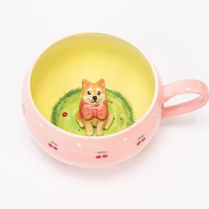 Puede incluir: Una taza de cerámica rosa con una figura de Shiba Inu en el interior. El interior de la taza es amarillo, con un parche de hierba verde y el perro con una pajarita roja. El exterior está decorado con lunares blancos y diseños de cerezas.