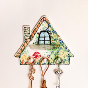 May include: A house-shaped ceramic wall hook rack featuring a painted garden scene with mushrooms, flowers, and a chimney. The piece includes a small integrated shelf below a window graphic and three white hooks holding various metal keychains.