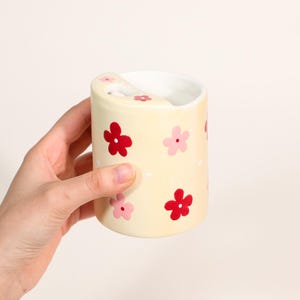 May include: A ceramic toothbrush holder with a light yellow background and red and pink flower designs. The holder has a curved top with a slot for toothbrushes. The interior is white.