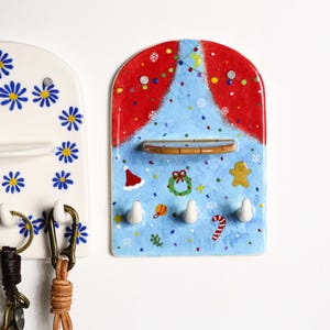Puede incluir: Un llavero de pared de cer&aacute;mica azul con tem&aacute;tica navide&ntilde;a. Presenta un dise&ntilde;o de cortina roja en la parte superior, una peque&ntilde;a repisa y cuatro ganchos blancos. El dise&ntilde;o incluye un gorro de Pap&aacute; Noel, una corona, un hombre de jengibre y un bast&oacute;n de caramelo.
