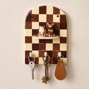 May include: An arched wall-mounted key holder featuring a brown and cream checkerboard pattern. It includes a small horizontal shelf displaying two miniature cat figurines and three white hooks at the bottom, each holding a set of metal keys.