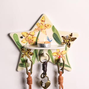 Puede incluir: Un llavero de cerámica en forma de estrella con un diseño floral con orquídeas amarillas y marrones y hojas verdes. Tiene un pequeño estante que sostiene una vela de té encendida y tres ganchos para llaves. Los llaveros son de cuero marrón.