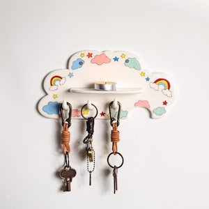 May include: A whimsical cloud-shaped key holder with a shelf for a small candle. The white ceramic holder is decorated with colorful rainbows, stars, and clouds. Three keychains with leather and metal accents hang below.