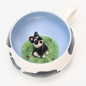 May include: A ceramic mug with a black and white cow pattern on the outside. Inside, a light blue sky design surrounds a green grassy area with a small dog figurine wearing a black jacket. The mug has a white handle.