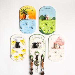 May include: Set of five ceramic key holders with dog figurines and the word "Welcome" at the top. Each holder features a different scene, including a beach, a field, and a floral design. Each has three hooks.