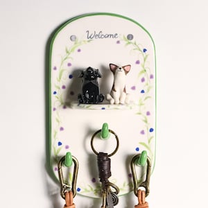 May include: A white ceramic key holder with a green border and the word "Welcome". It features two dog figurines and three green hooks. The key holder is decorated with purple and green floral accents.