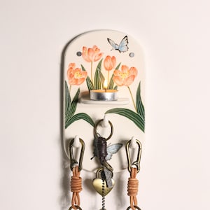 May include: Cream-colored key holder with a floral and butterfly design. Features a small candle holder with a lit candle, three key hooks, and leather keychains. The artwork includes orange tulips and a blue butterfly.