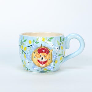 Puede incluir: Taza de cerámica azul claro con un diseño caprichoso. La taza presenta un cachorro en 3D con un sombrero y una bufanda rojos, sobre un fondo de limones, hojas y flores. La taza tiene un asa curva y un interior color crema.