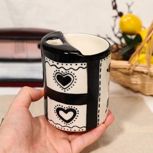 Pode incluir: Uma caneca de cerâmica preta e branca com um design de coração. A caneca tem formato retangular com alça e interior dividido. O design apresenta corações pretos em fundo branco, com borda preta e detalhes pontilhados. A caneca é segurada em uma mão.