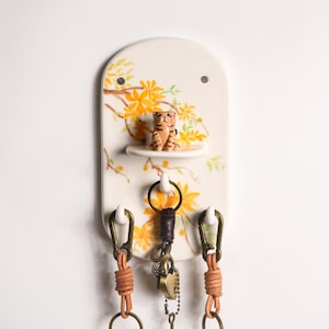 Könnte beinhalten: Ein keramischer Schlüsselhalter mit einem floralen Design in Gelb und Orange. Er verfügt über ein kleines Regal mit einer Tigerfigur und drei Haken mit Leder- und Metallakzenten. Der Schlüsselhalter ist an einer weißen Wand befestigt.