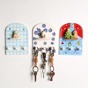 May include: Three ceramic key holders with decorative designs. One is blue gingham with a dog figurine, another has blue flowers and a cat figurine, and the third is Christmas-themed with a tree and dog figurine. Each has hooks for keys.
