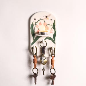 May include: A ceramic key holder with a floral design, a small shelf holding a lit candle, and three key hooks. The key holder features a bee and pink flowers. The keys are attached to leather straps.