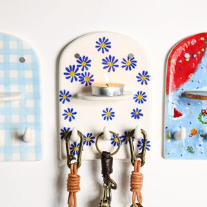 Puede incluir: Un juego de tres llaveros de cer&aacute;mica. El soporte central es blanco con dise&ntilde;os de margaritas azules, una peque&ntilde;a repisa con una vela encendida y tres ganchos con llaveros de cuero. Los otros dos soportes tienen un dise&ntilde;o de cuadros azules y un dise&ntilde;o festivo.