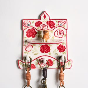May include: A ceramic key holder featuring a red rose design and a small dog figurine. The key holder has three hooks for keys and is decorated with red roses and green leaves.
