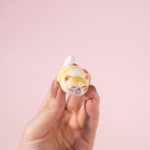 May include: A small, ceramic cat figurine in a relaxed pose. The cat is primarily orange and white with a smiling face and closed eyes. The figurine is held against a soft pink background.