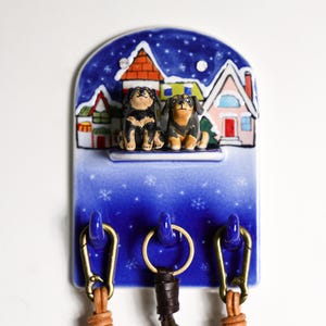 May include: A blue ceramic key holder featuring two dog figurines in front of a winter village scene. The key holder has three hooks with leather straps and gold-colored clasps. The key holder is approximately 8 inches tall.