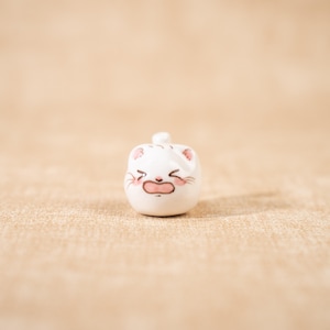Könnte beinhalten: Kleine, weiße Keramik-Katzenfigur mit einem betrübten Gesichtsausdruck. Die Katze hat rosa Akzente an Ohren und Wangen und einen kleinen, runden Körper. Die Figur ist ein dekoratives Objekt.