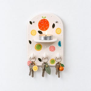 May include: An arched cream-coloured ceramic wall organiser featuring a small shelf holding a lit tea light candle. The surface is decorated with painted citrus fruit motifs and white dots. Three hooks at the bottom hold metal keys with charms.