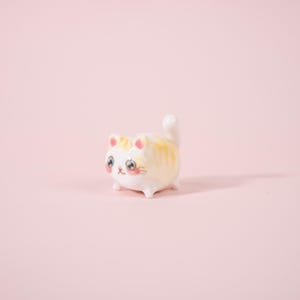 May include: A ceramic cat figurine with a white base and yellow and orange stripes. The cat has large, expressive eyes, pink cheeks, and a small, upturned tail. The figurine is on a pink background.