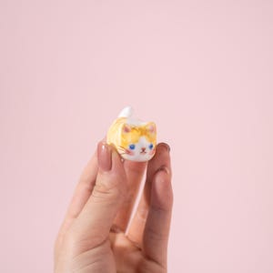 Könnte beinhalten: Kleine Keramik-Katzenfigur mit gelb-orangefarbenem Körper, weißen Akzenten und blauen Augen. Die Katze liegt mit erhobenem Schwanz. Die Figur wird in einer Hand vor rosa Hintergrund gehalten.
