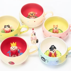 Könnte beinhalten: Eine Sammlung von Keramikbechern mit farbenfrohen Innen- und Außendesigns. Jeder Becher zeigt eine andere Tierfigur im Inneren, darunter ein Hund, eine Katze und ein Piratenhund. Die Becher haben Henkel und stehen vor einem weißen Hintergrund.