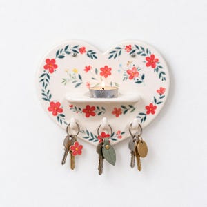 May include: A heart-shaped ceramic wall organiser featuring a cream background with red floral and green leaf patterns. It includes a small central shelf holding a lit tea light candle and three hooks at the bottom, each holding a set of keys.