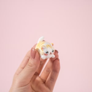 May include: A small, ceramic cat figurine with a white base and orange stripes. The cat has a cute, cartoonish design with blue eyes, pink cheeks, and a small white tail. The figurine is held in a hand against a pink background.