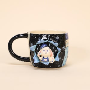Pode incluir: Uma caneca de cerâmica com exterior preto decorada com flocos de neve brancos e um design de patchwork azul e branco. A caneca apresenta um coelho esculpido em 3D usando um chapéu azul e macacão, segurando um pincel e uma paleta.