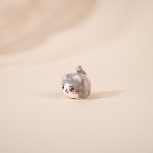 Op de afbeelding: Een kleine keramische kattenfiguur, voornamelijk grijs en wit, met grote, expressieve ogen en een kleine staart. De kat heeft roze wangen. Het beeldje staat tegen een neutrale achtergrond.