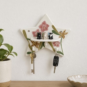 Puede incluir: Un soporte para llaves de pared de cerámica en forma de estrella con un patrón floral de orquídeas pintadas. La pieza incluye un pequeño estante horizontal con tres figuras de gatos en miniatura y dos ganchos blancos con llaves.