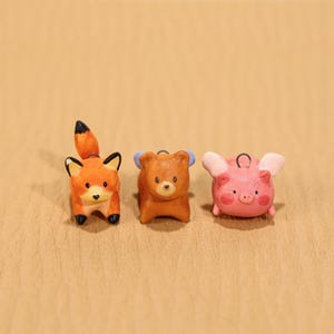 May include: Three small animal figurines: a fox, a bear, and a pig. The fox is orange with a black-tipped tail. The bear is brown with blue accents. The pig is pink with small wings. All are on a tan surface.
