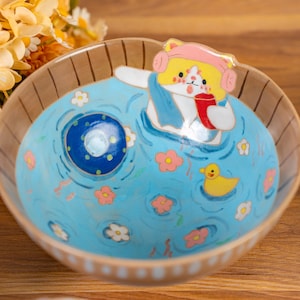 Può includere: Una ciotola in ceramica dal design stravagante. L'interno presenta una scena di piscina azzurra con un gatto dei cartoni animati con un cappello rosa e una tazza rossa. Fiori rosa e un'anatra di gomma gialla galleggiano nell'acqua. Il bordo della ciotola è marrone con strisce verticali.