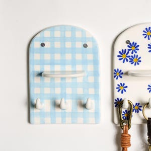 Puede incluir: Un llavero de pared con estampado de cuadros vichy azul y blanco, tres ganchos y una peque&ntilde;a repisa. El llavero de cer&aacute;mica tiene una parte superior redondeada con dos peque&ntilde;os puntos negros. El dise&ntilde;o evoca una est&eacute;tica alegre de estilo campestre.