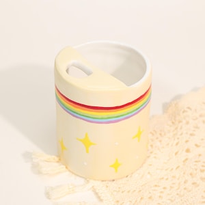 Pode incluir: Um porta-escovas de dentes cilíndrico de cerâmica creme com uma faixa de arco-íris e detalhes de estrelas amarelas. O suporte tem uma alça embutida e um interior branco. O design é caprichoso e adequado para um banheiro.