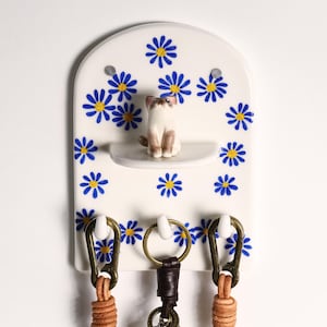 May include: A white ceramic key holder with a rounded top, decorated with blue daisy patterns. It features a small shelf with a cat figurine and three hooks with leather keychains. The key holder is mounted on a white wall.